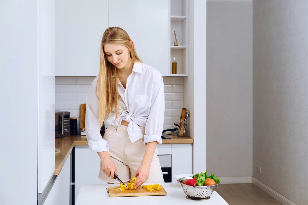 A wide shot of Sarah Miller in her kitchen, focused on chopping a yellow pepper on a cutting board with fresh vegetables nearby.
