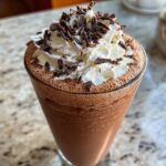 Close-up of a tall glass filled with rich, blended frozen hot chocolate, topped with whipped cream and chocolate shavings.