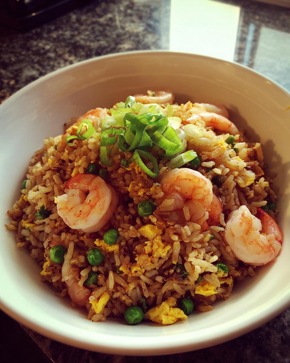 A close-up of a white bowl filled with flavorful shrimp fried rice, topped with pink shrimp, green peas, scrambled egg, and sliced scallions.
