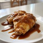 Two golden-brown apple dumpling pastries drizzled generously with rich caramel sauce on a white plate.