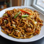 A close-up of a generous serving of homemade chicken chow mein recipe with noodles, chicken pieces, carrots, and green onions.