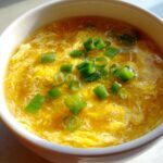 A close-up of a white bowl filled with bright yellow egg drop soup, garnished with fresh green scallions.