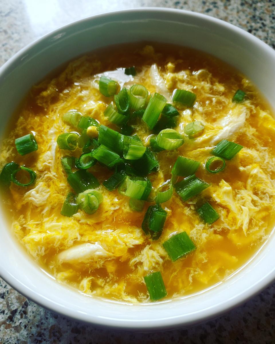 A close-up shot of a bowl of hot egg drop soup, featuring wispy yellow egg ribbons topped with freshly sliced green onions.