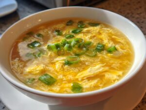 A close-up shot of a white bowl filled with steaming, savory egg drop soup garnished with fresh green scallions.