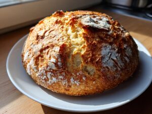 A freshly baked, round loaf of amazing italian bread with a deeply golden, cracked crust dusted with flour, sitting on a white plate.