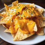 Close-up of tortilla chips generously covered in bright orange nacho cheese sauce and sprinkled with parsley.