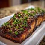 A close-up of a perfectly cooked fillet of teriyaki salmon, heavily glazed and topped with sesame seeds and chopped green onions.