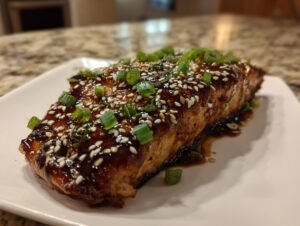 A perfectly cooked fillet of teriyaki salmon glazed with a dark sauce, topped with white sesame seeds and chopped green onions.