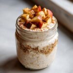 Close-up of apple cinnamon overnight oats in a glass jar, topped with yogurt, diced red apples, and cinnamon.