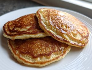 Close-up of three golden brown banana oatmeal pancakes stacked on a white plate, glistening with syrup.