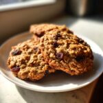 Three golden brown oatmeal raisin cookies stacked slightly on a white plate, catching bright sunlight.