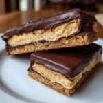 Two layered bars of buckeye graham crackers stacked on a white plate, showing peanut butter filling and chocolate topping.