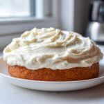 A round cake generously topped with thick, swirled cream cheese frosting, sitting on a white plate.