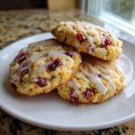 Three chewy cranberry orange cookies stacked on a white plate, drizzled with white icing.