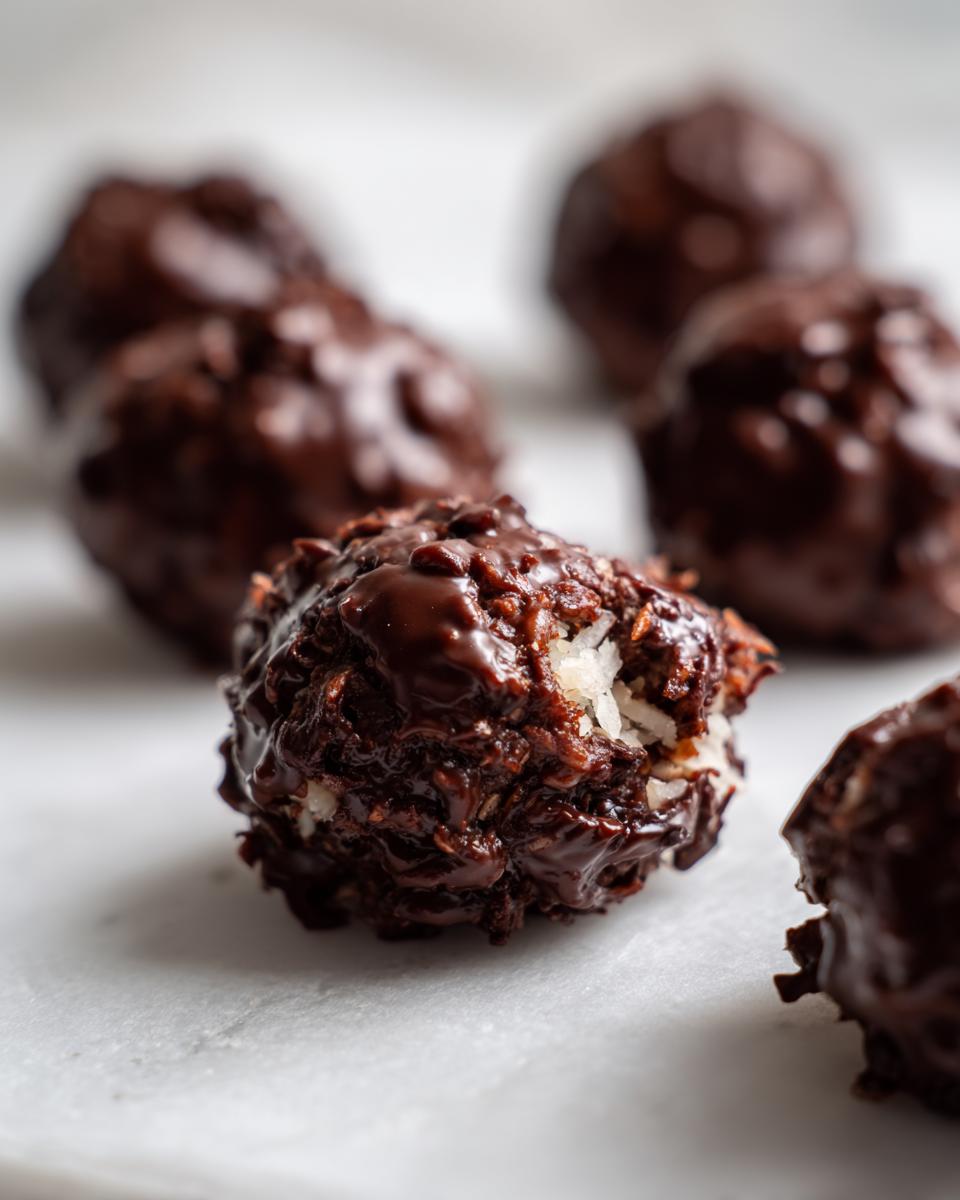 A close-up of a rich, dark chocolate-covered coconut balls, showing shredded coconut texture inside a bite taken out.