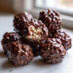 A stack of rich, chocolate-covered coconut balls, with one bitten open revealing the shredded coconut interior.
