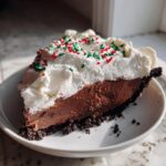 A decadent slice of Christmas chocolate cream pie with a thick chocolate filling, whipped cream, and red and green holiday sprinkles.