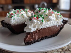 A close-up of a slice of Christmas chocolate cream pie featuring a dark crust, rich filling, and whipped cream topped with red and green sprinkles.
