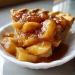 A close-up of a slice of pie topped with generous spoonfuls of glistening, spiced apple pie filling.