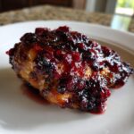 A close-up photo of a piece of baked cranberry chicken covered in a thick, glossy, dark red cranberry glaze.