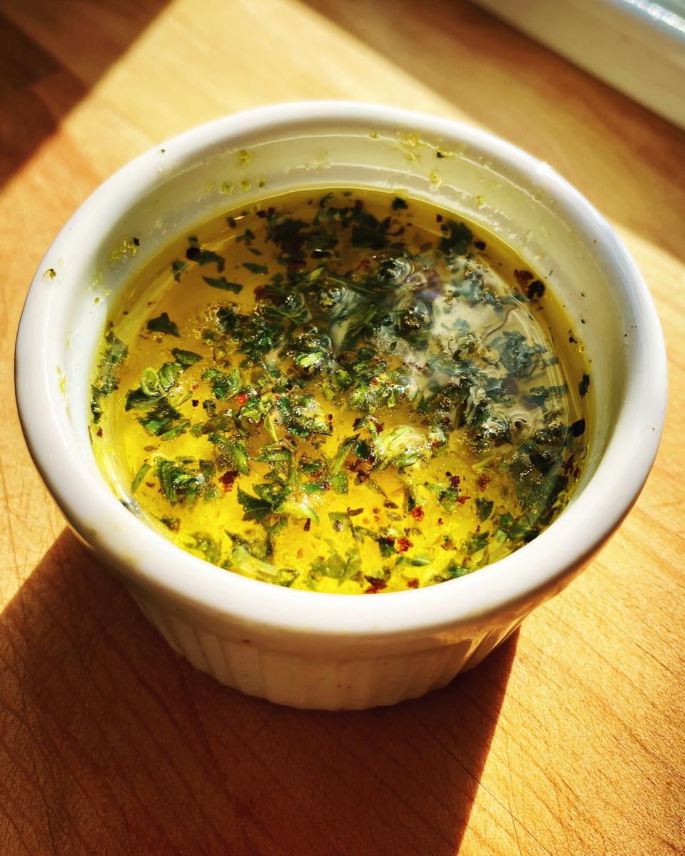 Close-up of melted, bright yellow cowboy butter dipping sauce flecked with green herbs and red pepper flakes in a white ramekin.