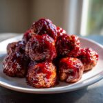 A close-up of several glossy cranberry meatballs piled high on a white plate, coated in a rich, dark red sauce.