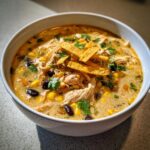 Close-up of a bowl of creamy chicken tortilla soup, filled with shredded chicken, corn, black beans, and topped with tortilla strips and cilantro.