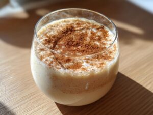 Close-up of a glass filled with creamy, light-colored coquito, heavily dusted with ground cinnamon.