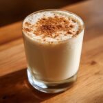 A glass of creamy coquito topped with a generous dusting of ground cinnamon, sitting on a wooden surface.