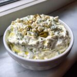 A white bowl filled with creamy dirty martini dip, topped generously with crumbled blue cheese and sliced green olives.