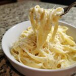 A fork lifting fettuccine noodles coated in creamy homemade alfredo sauce, topped with grated Parmesan cheese.