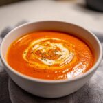 Close-up of a bowl of rich, orange tomato bisque topped with a swirl of cream and herbs.