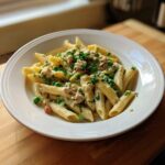 A close-up of creamy tuna pasta featuring penne, green peas, and fresh parsley garnish.