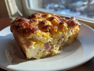 A square slice of baked Croissant Breakfast Casserole with ham and cheese, featuring a golden-brown, crusty top.