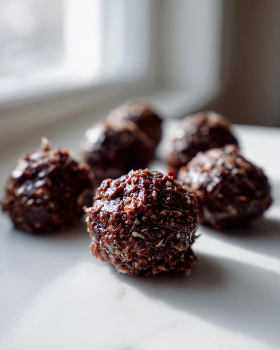 A close-up, low-light photo of several rich, dark chocolate coconut balls resting on a white surface.