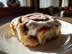 A single, fluffy cinnamon roll generously topped with white cream cheese frosting, sitting on a white plate.