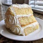 A thick, three-layer slice of fluffy vanilla cake with white buttercream frosting, served on a white plate.