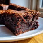 A close-up, macro shot of a rich, dark, fudgy piece of sweet potato brownies with a crackly top, resting on a white plate.