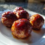 Four glistening Grape Jelly Meatballs covered in a thick, dark red glaze, resting on a white plate.