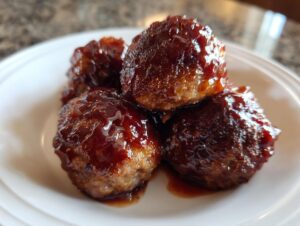 Close-up of four glistening Grape Jelly Meatballs covered in a thick, dark, sweet glaze, served on a white plate.