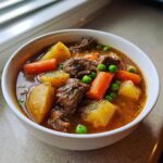 Close-up of a rich vegetable beef soup in a white bowl, featuring tender beef chunks, potatoes, carrots, and bright green peas.