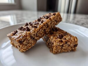 Two homemade chocolate chip protein bars stacked slightly on a white plate, showing oat texture.