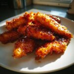 Close-up of crispy, glazed hot honey chicken tenders piled on a white plate, glistening under bright light.