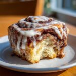 A delicious, gooey cinnamon roll topped with white icing, sitting on a small plate with one bite missing.