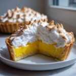 Close-up of a lemon meringue tart cut in half, showing bright yellow filling and toasted meringue peaks.