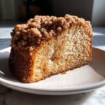 A close-up slice of moist coffee cake featuring a thick, sugary cinnamon crumb topping, beautifully lit by sunlight.