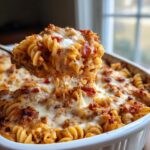A spoonful of cheesy, saucy pizza pasta being lifted from a white baking dish.