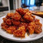 Close-up of crispy pieces of orange chicken recipe coated in a thick, glossy orange sauce and sprinkled with sesame seeds.
