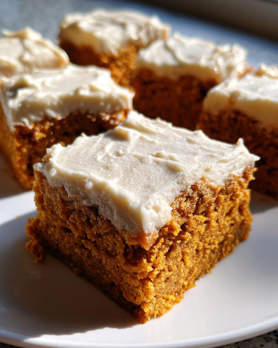 A close-up of a square slice of moist pumpkin bars with cream cheese frosting on a white plate.
