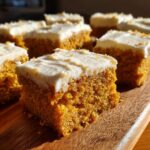 A close-up of a square piece of moist pumpkin bars with cream cheese frosting, sitting on a wooden board.